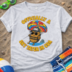 Gray t-shirt with a humorous graphic design of a potato skydiving, placed on sand with beach accessories.